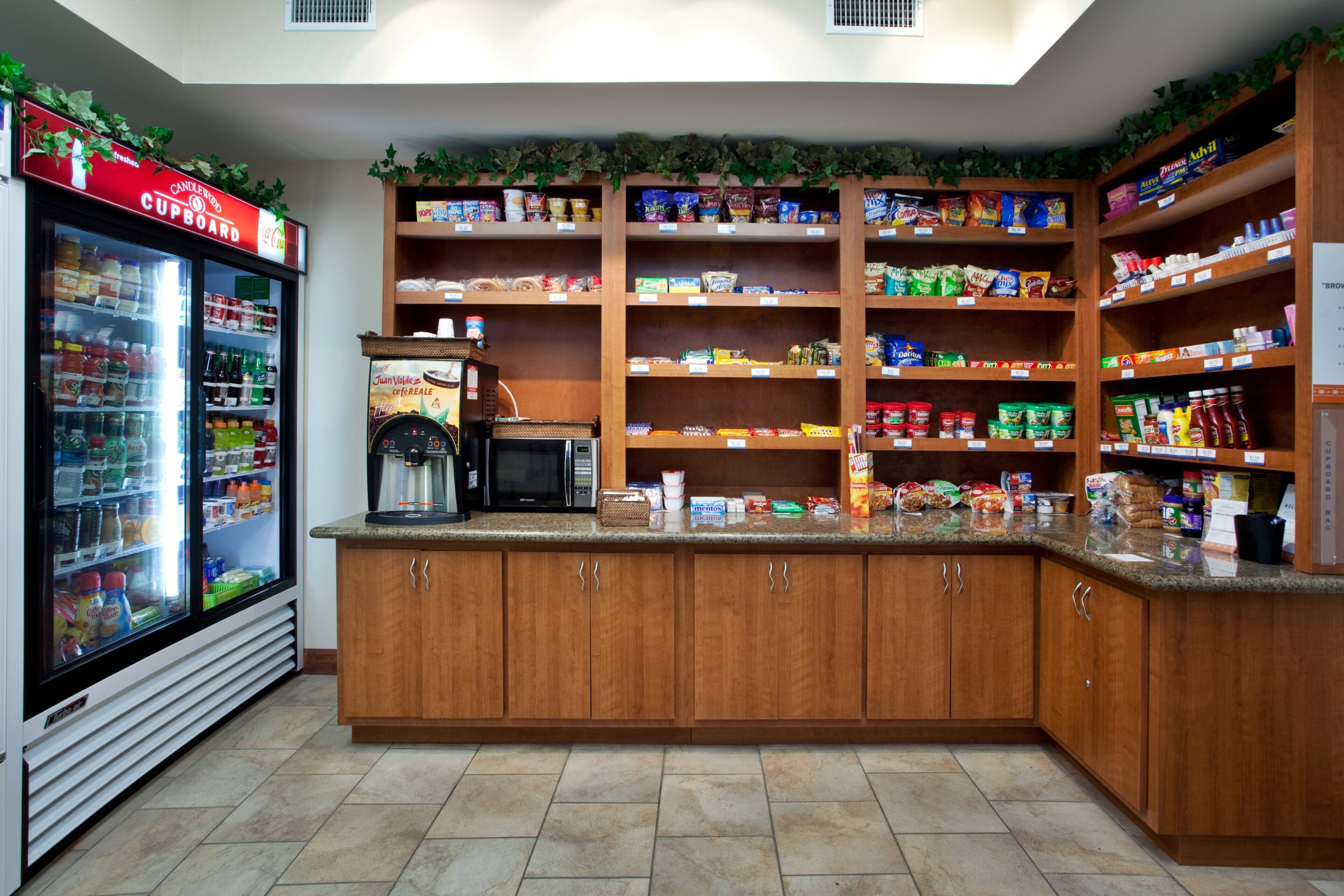 A self-service food area with a refrigerated drink cooler, a coffee machine, and shelves stocked with snacks and condiments.
