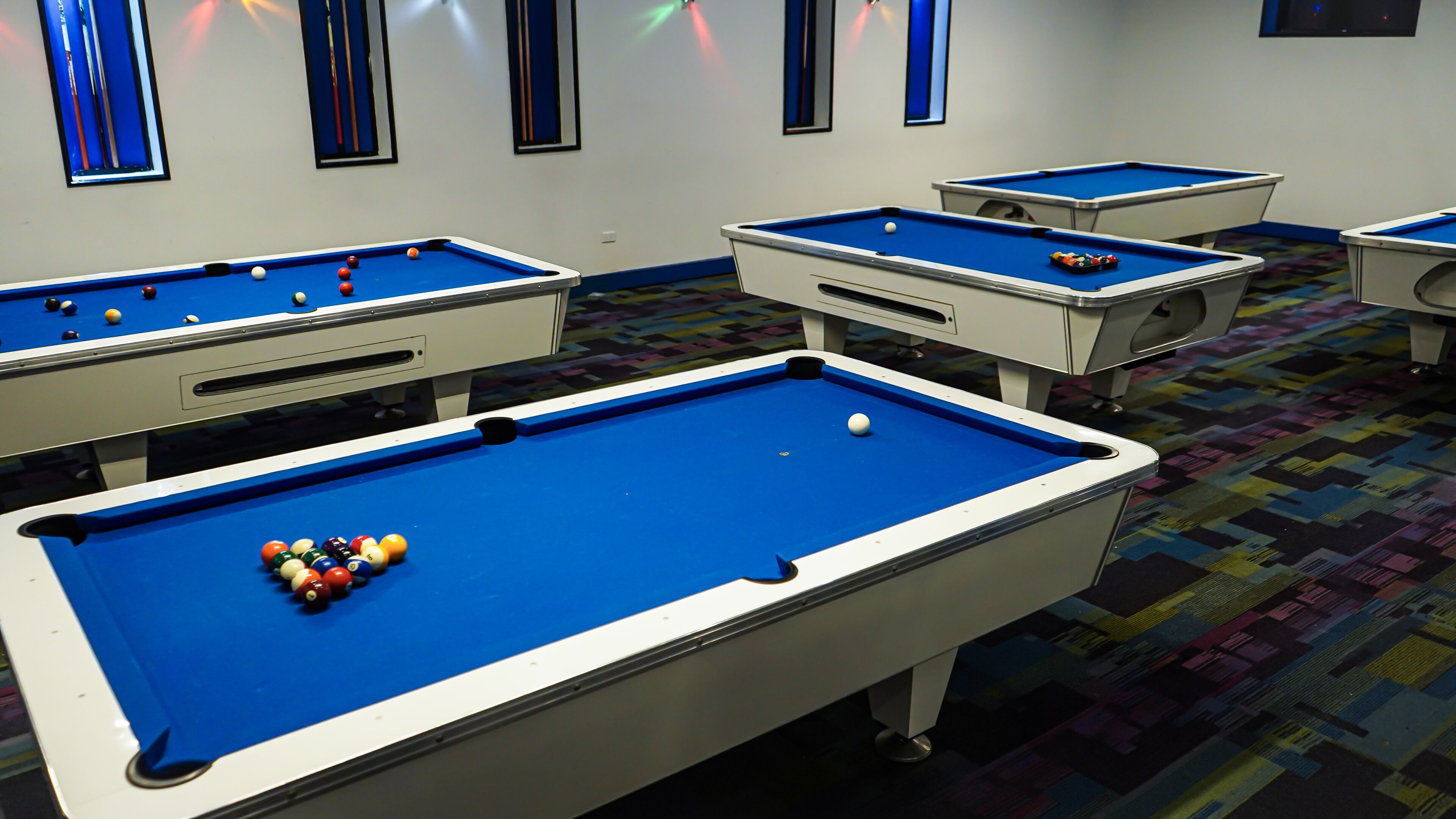 Una sala de billar con múltiples mesas, cada una con superficie de fieltro azul y bordes blancos.