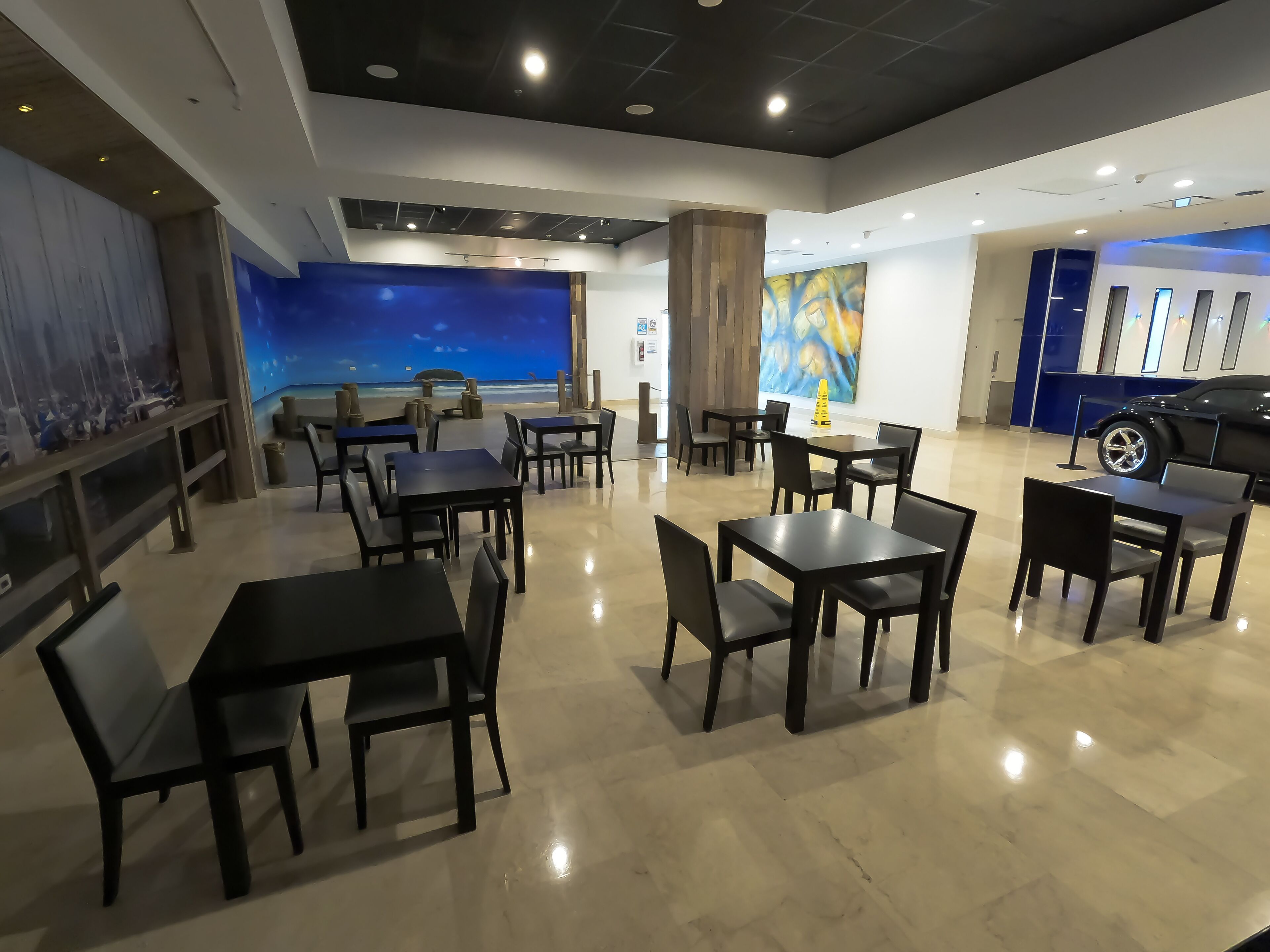 Un restaurant moderne offrant une vue sur l’océan, avec de nombreuses tables et chaises, un grand tableau et une exposition de voitures.