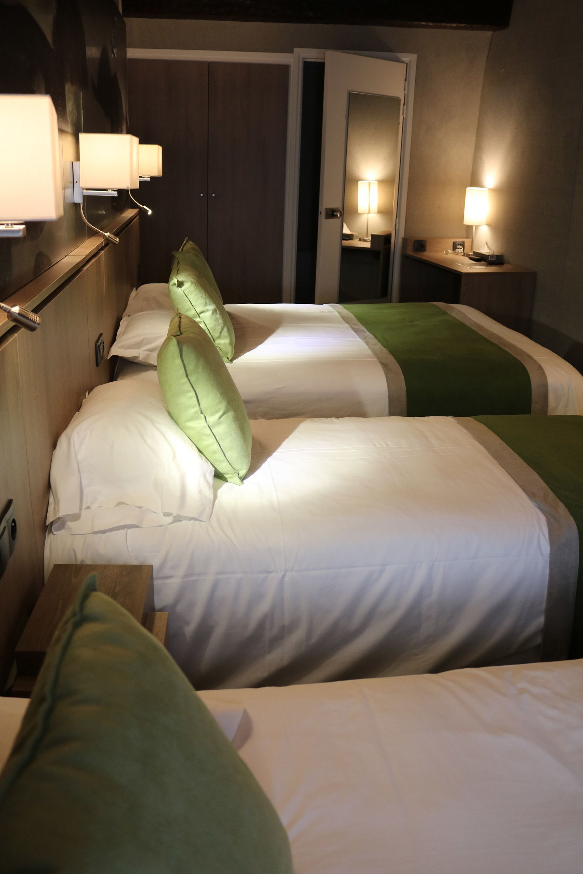 A hotel room with two beds, each with white bedding and green pillows. There are bedside lamps and a mirror above the beds.