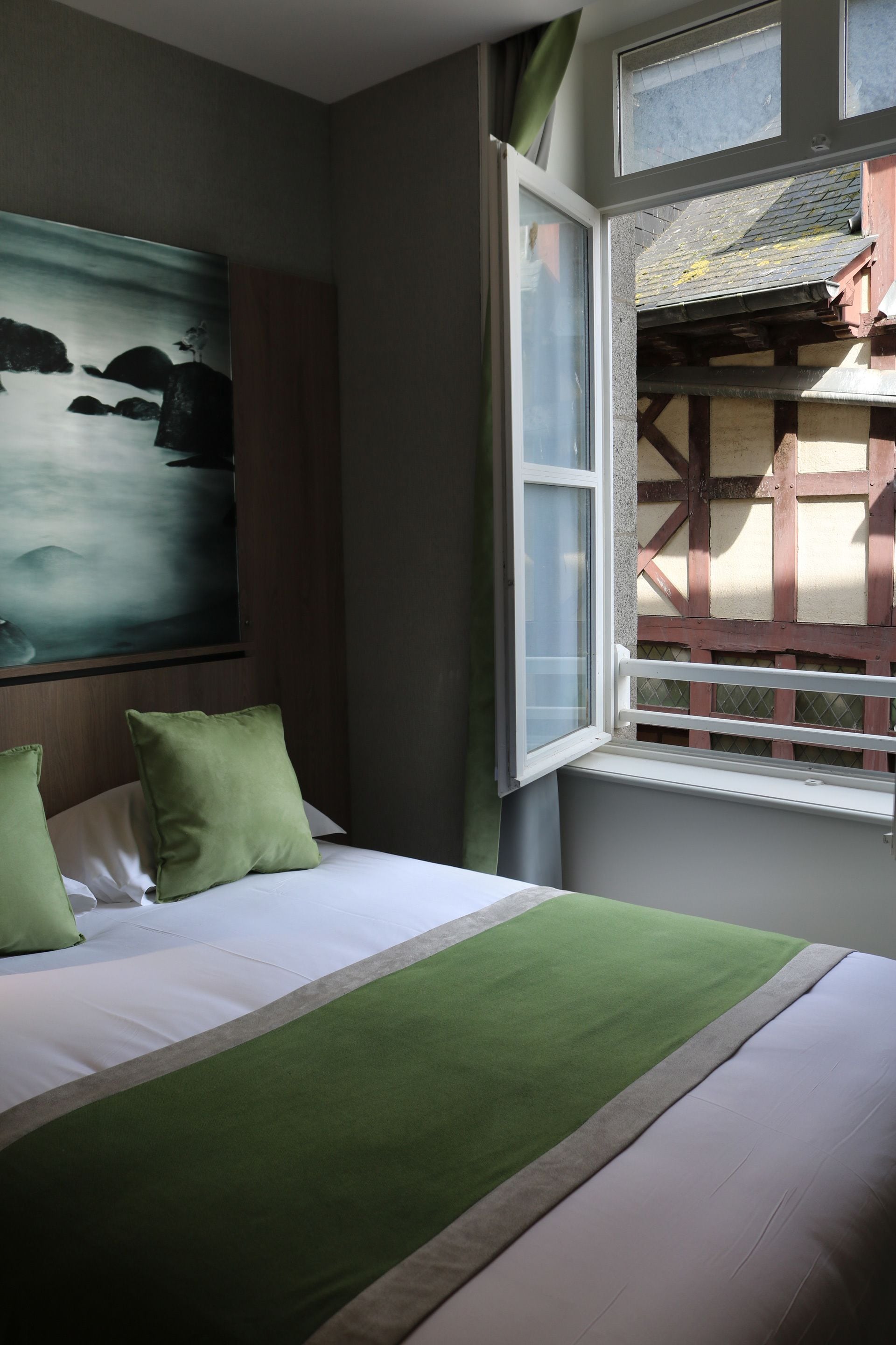 A hotel room with a bed, a painting, and a window with a view of a building.