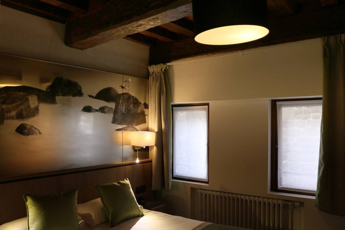 Ein Hotelzimmer mit einem Bett, zwei Kissen, einem Nachttisch mit Lampe, einer Wanduhr und zwei mit Vorhängen versehenen Fenstern.