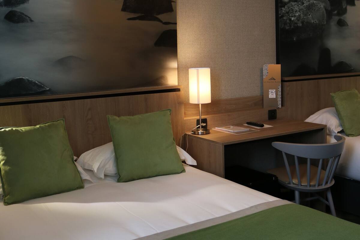 Ein Hotelzimmer mit einem Bett, grünen Kissen, einem Schreibtisch mit Lampe, einem Sessel und zwei großen Bildern an der Wand.