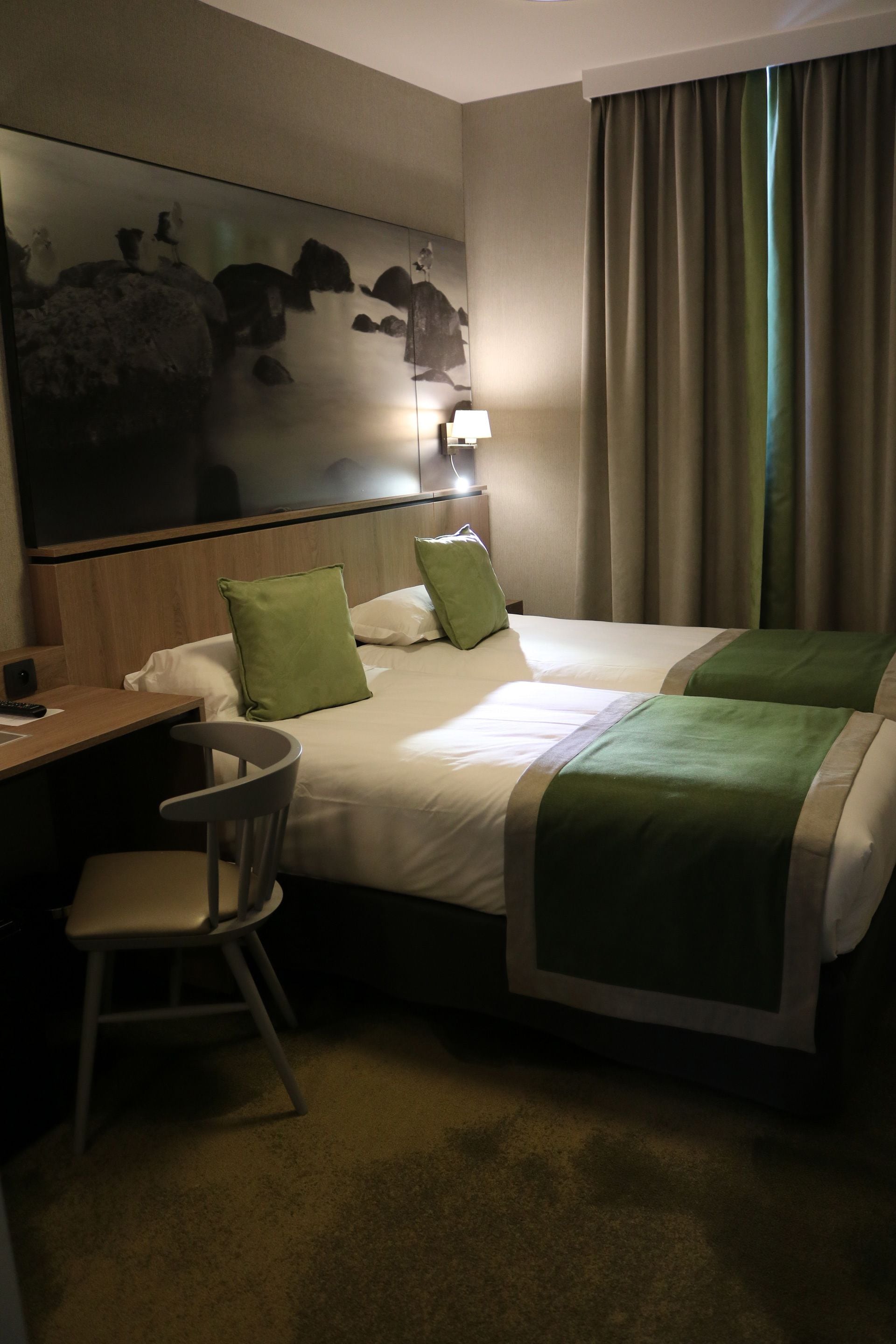 Ein Hotelzimmer mit zwei Betten, einem Schreibtisch, einem Stuhl, einem Bild an der Wand und einem Fenster mit Vorhängen.