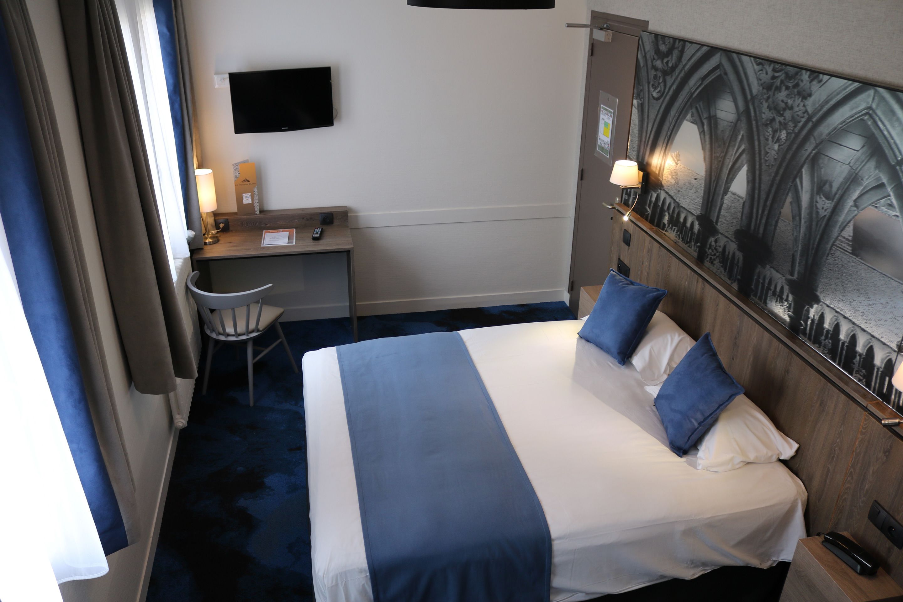 A hotel room with a bed, a desk, a chair, a TV, and a painting on the wall.