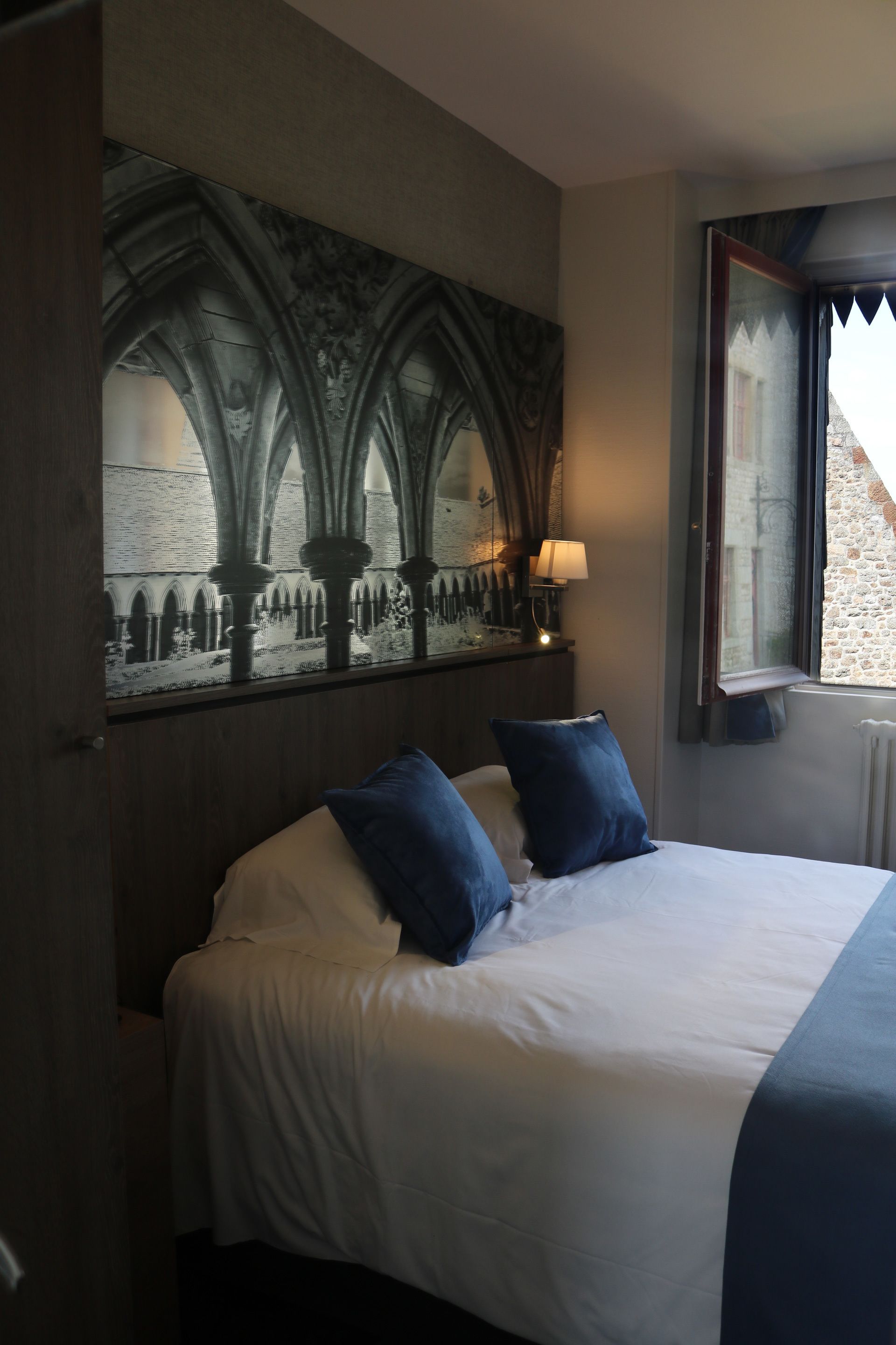 Une chambre avec un grand lit, deux oreillers, une table de chevet avec une lampe et une fenêtre donnant sur un bâtiment en pierre.