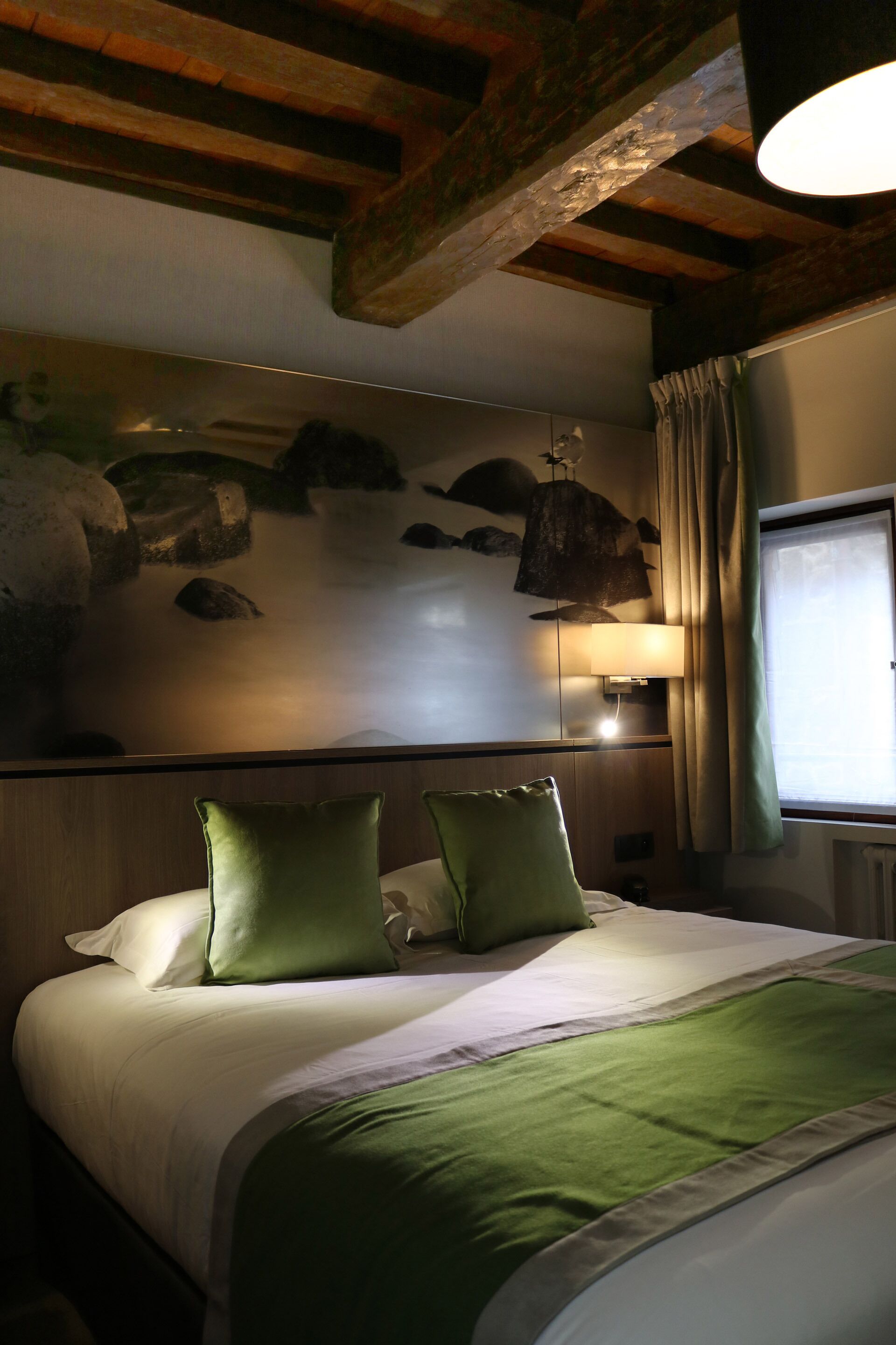 Una habitación de hotel con una cama, almohadas verdes, una lámpara de pared y una ventana con cortinas, además de una decoración mural de piedra.
