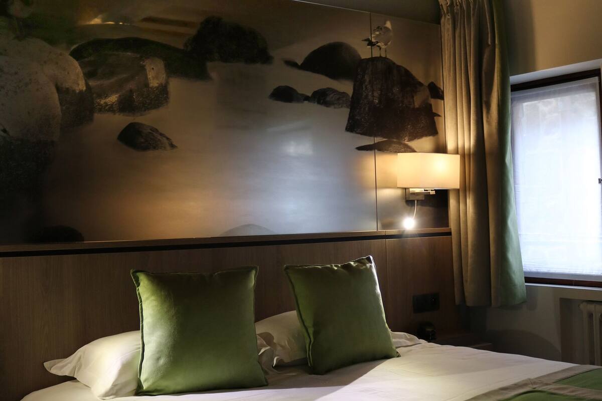 A hotel room with a bed, green pillows, a wall-mounted lamp, a window with curtains, and a stone wall decoration.