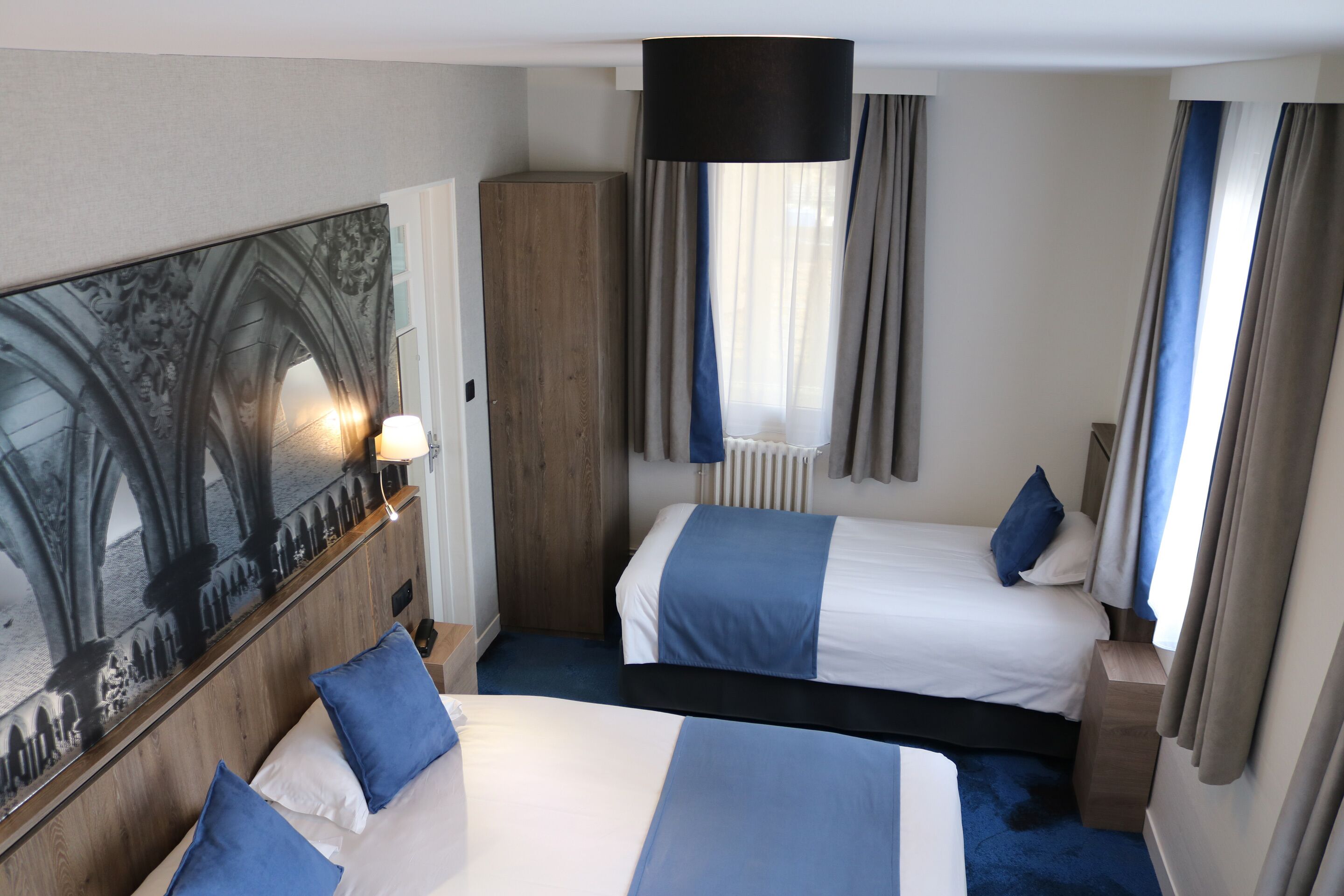 Habitación de hotel con dos camas, un cuadro grande, un armario de madera y una ventana con cortinas.