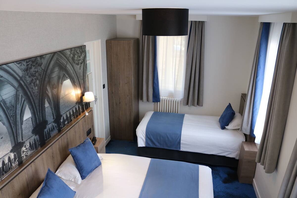 A hotel room with two beds, a large painting, a wooden wardrobe, and a window with curtains.