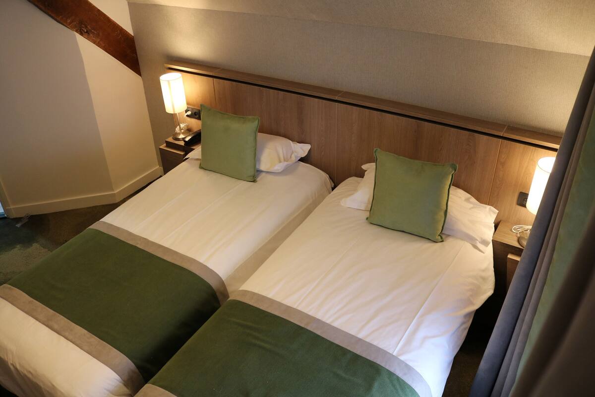 A hotel room with two beds, each with a green pillow and white linens. There are bedside tables with lamps and a wooden headboard.