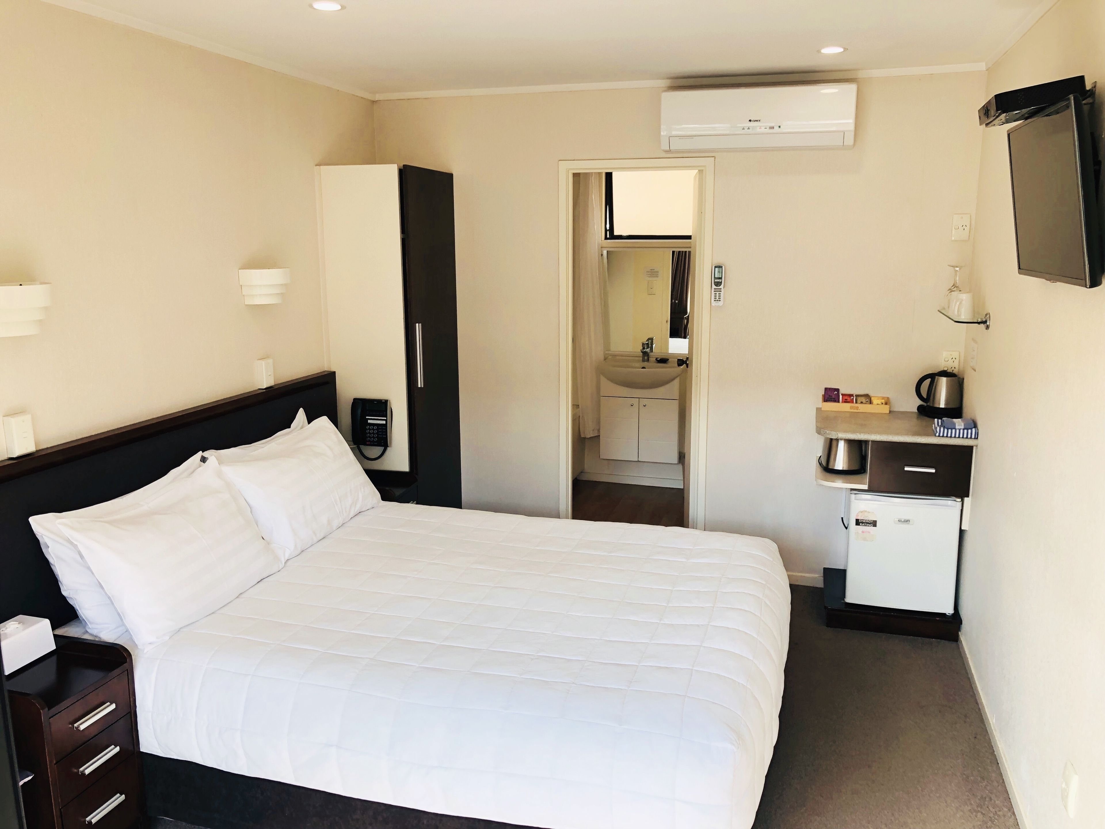 A hotel room with a bed, a small kitchenette, a bathroom, and a TV mounted on the wall.