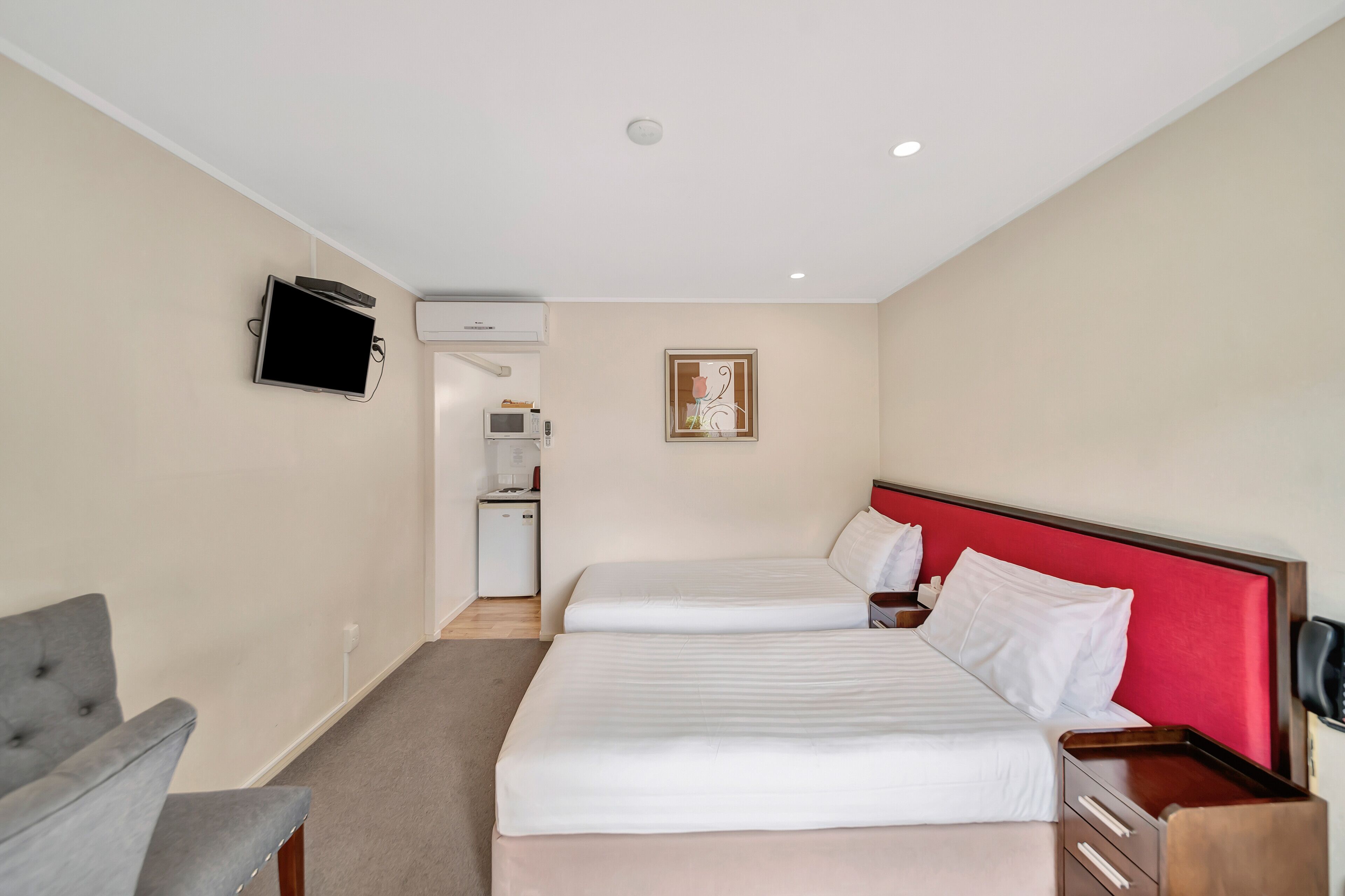 A hotel room with two beds, a TV mounted on the wall, a small kitchenette, and a desk with drawers.