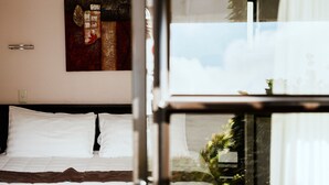 A hotel room with a bed, white pillows, a painting on the wall, and a view of greenery outside. - Tudor Court Motor Lodge (Auckland)