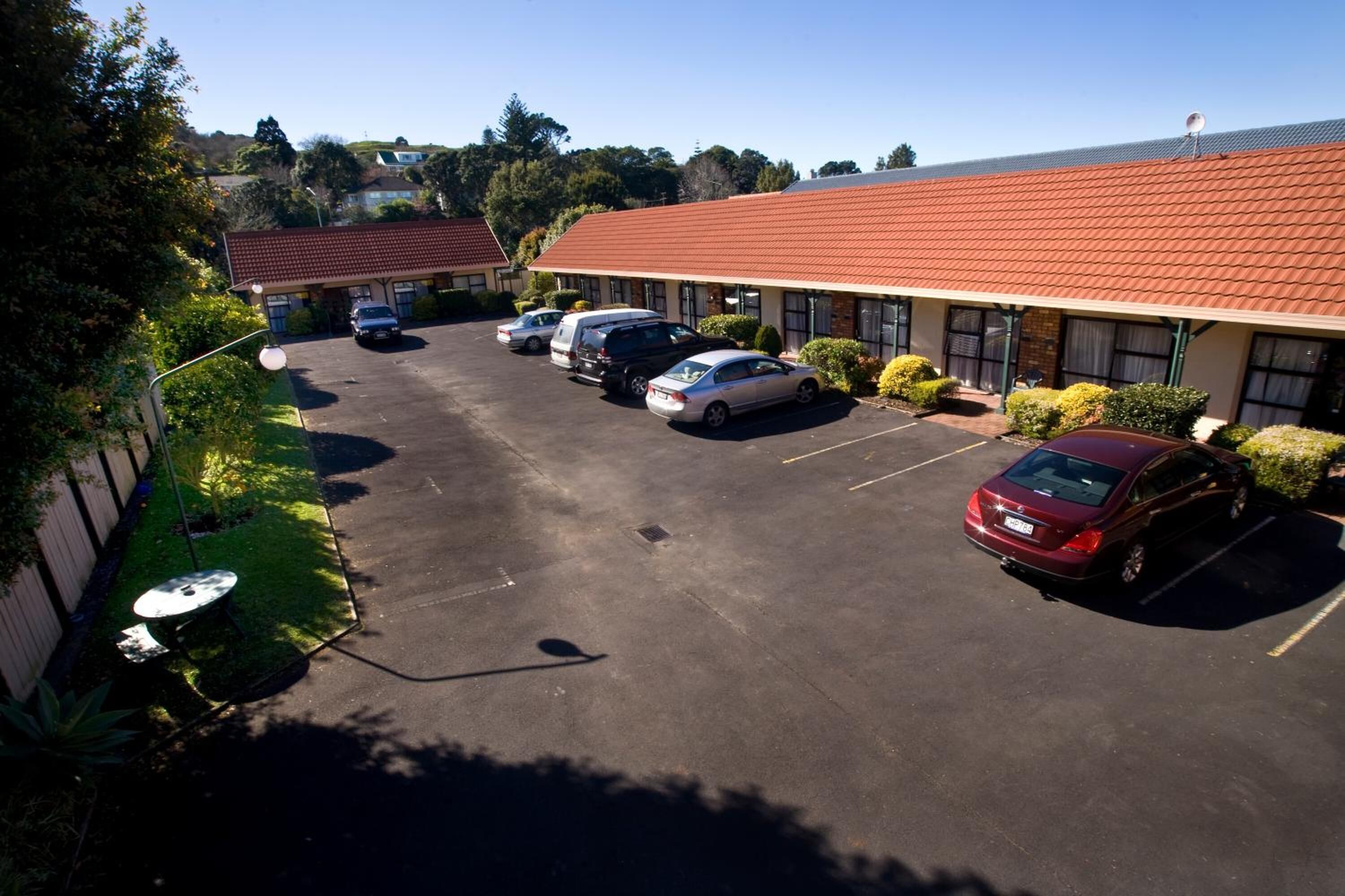 A motel with a red-tiled roof, a parking lot with several cars, and a small garden area.