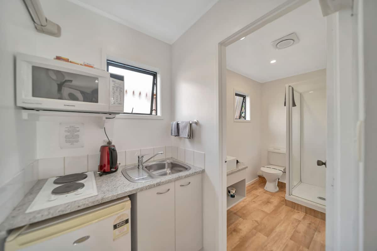 Cocina compacta con microondas, estufa, pileta y un ventanal pequeño. Junto a ella, un baño con inodoro, ducha y cabina de ducha con mampara de vidrio.