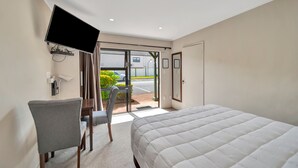 A hotel room with a bed, a desk, a chair, a TV mounted on the wall, and a sliding glass door leading to an outdoor area. - Tudor Court Motor Lodge (Auckland)