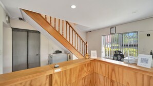 A reception area with a wooden staircase, a desk with a computer, a chair, and a window with a view of the outside. - Tudor Court Motor Lodge (Auckland)