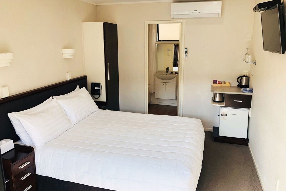 A hotel room with a bed, a desk with a phone, a small kitchenette, and a bathroom visible through an open door.