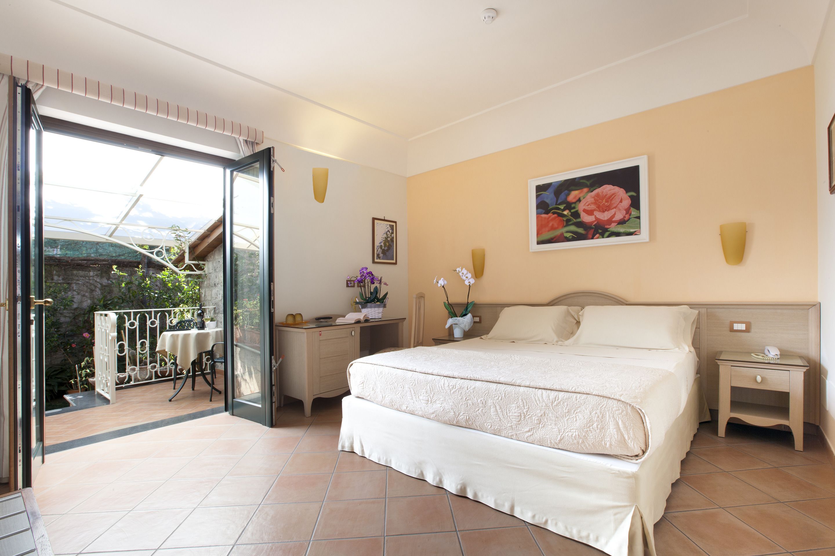Une chambre avec un grand lit, une table de chevet, un bureau avec une chaise et un balcon avec une table et des chaises.