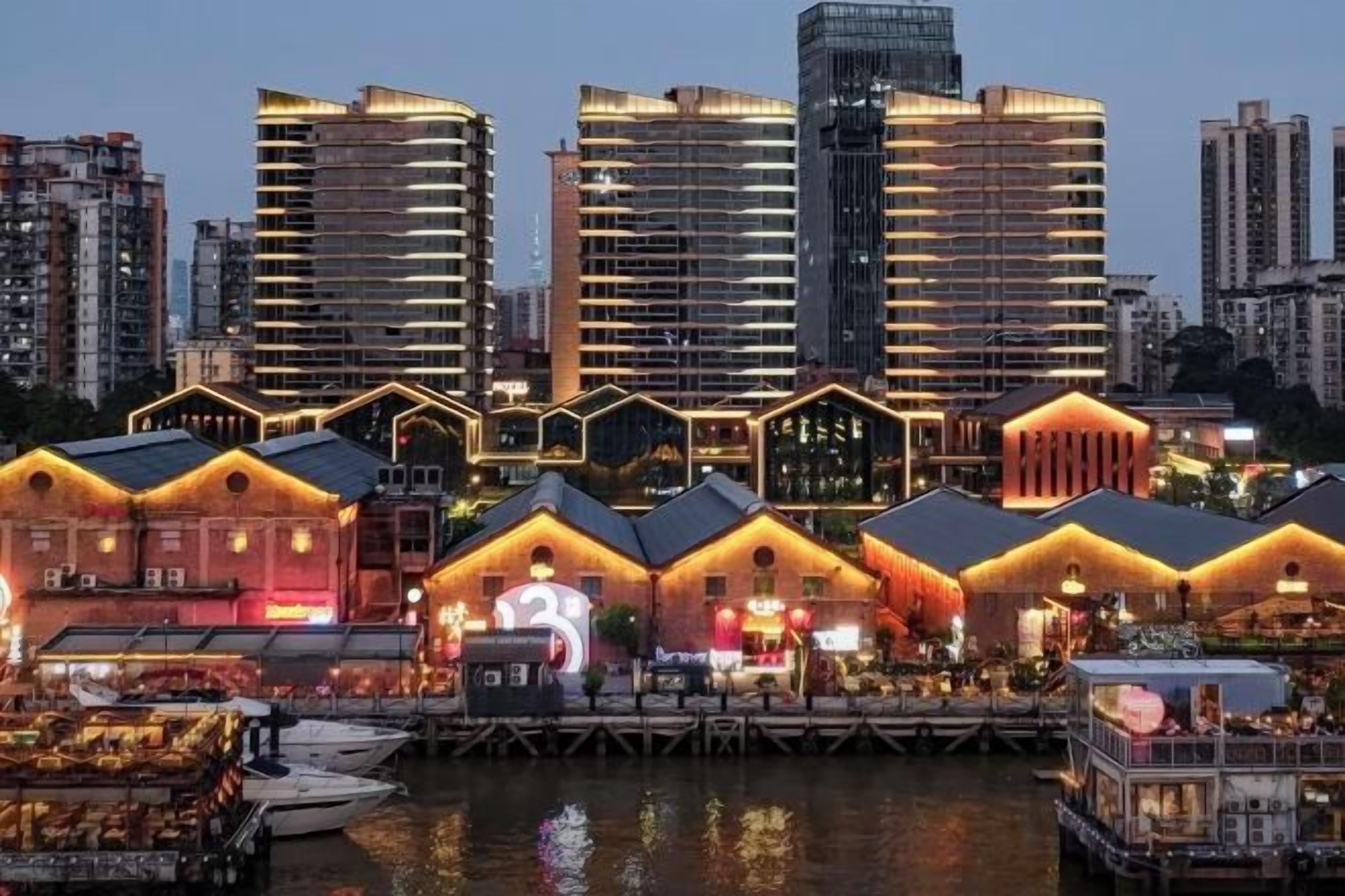 Un hotel frente al agua con varias construcciones, un muelle con barcos y un horizonte urbano al fondo.