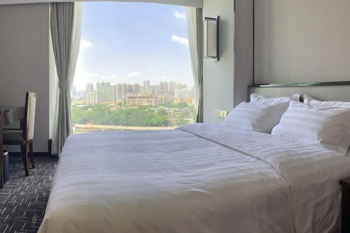 Quarto de hotel com uma cama grande, uma cadeira, uma TV e vista para a cidade pela janela.