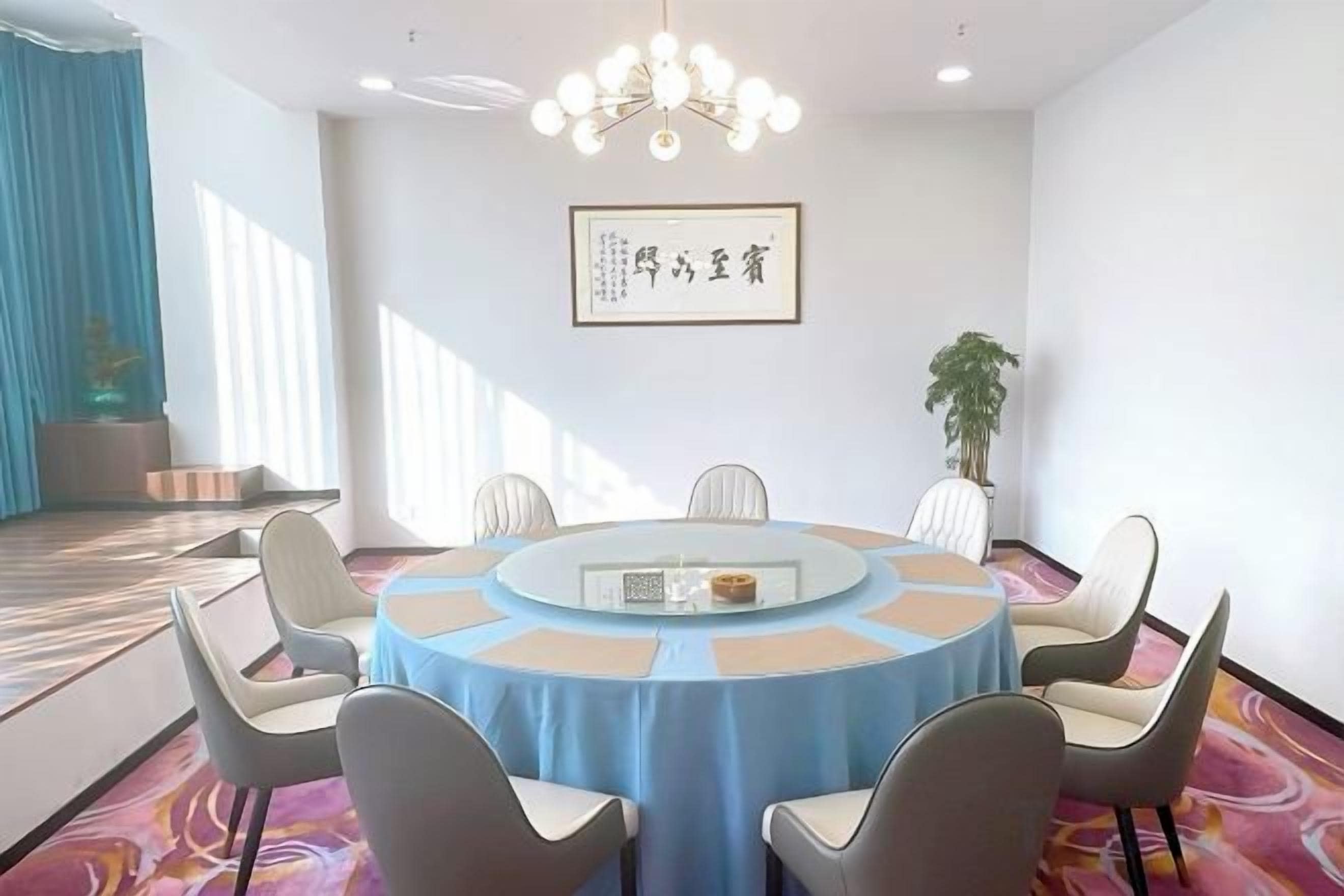 Un salón de conferencias con una mesa redonda, sillas, una araña y una obra de arte enmarcada en la pared.