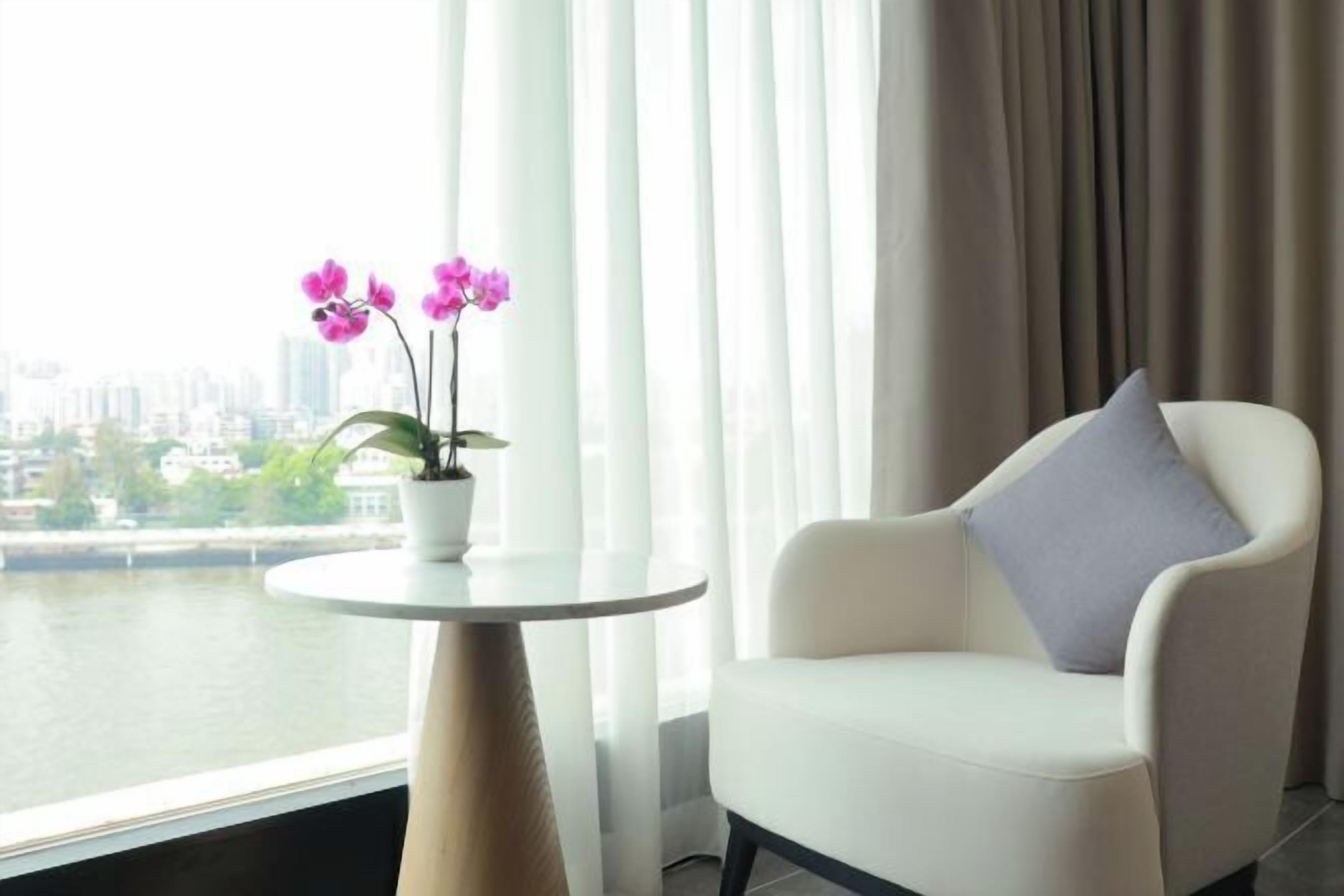 Una habitación moderna con un sillón blanco y una pequeña mesa con una orquídea en maceta. Se ve un paisaje urbano a través de la ventana.