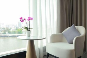 Quarto moderno com poltrona branca e pequena mesa com uma orquídea em vaso. Uma vista urbana é visível pela janela.