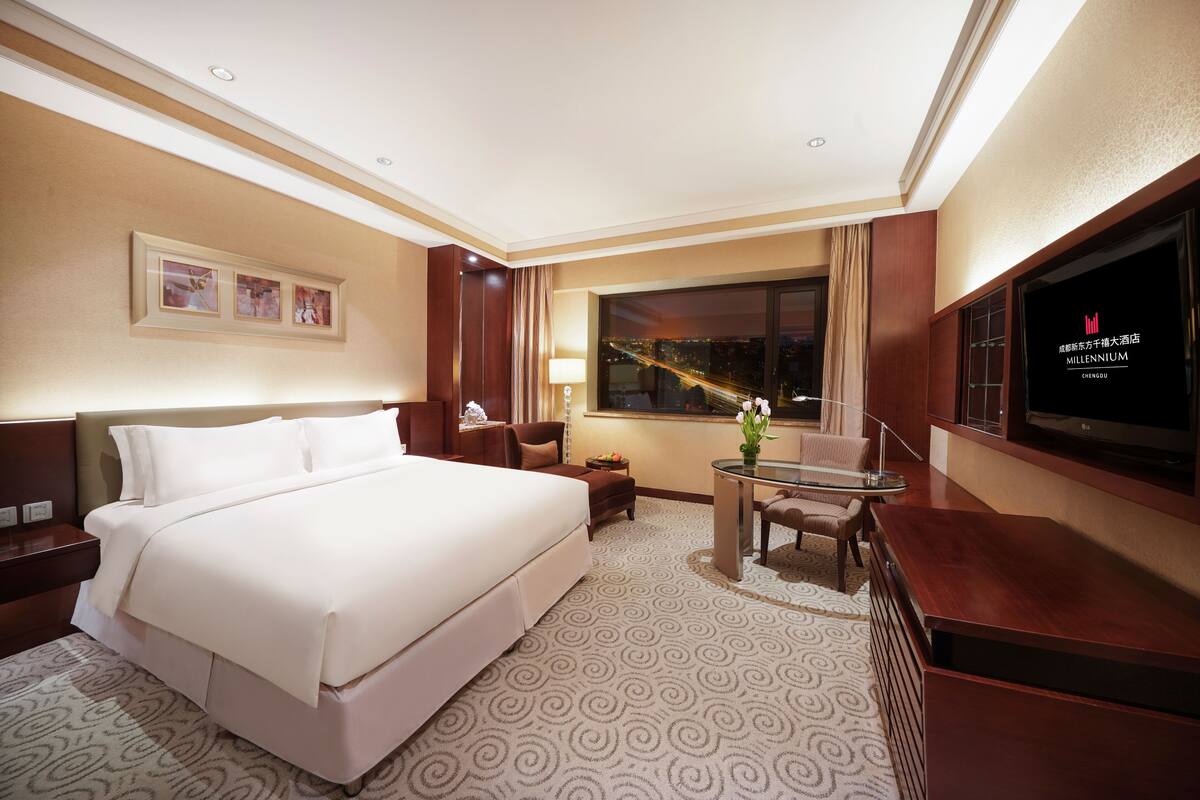 Habitación de hotel con una cama grande, un escritorio con silla, televisor y ventana con vista a la ciudad.