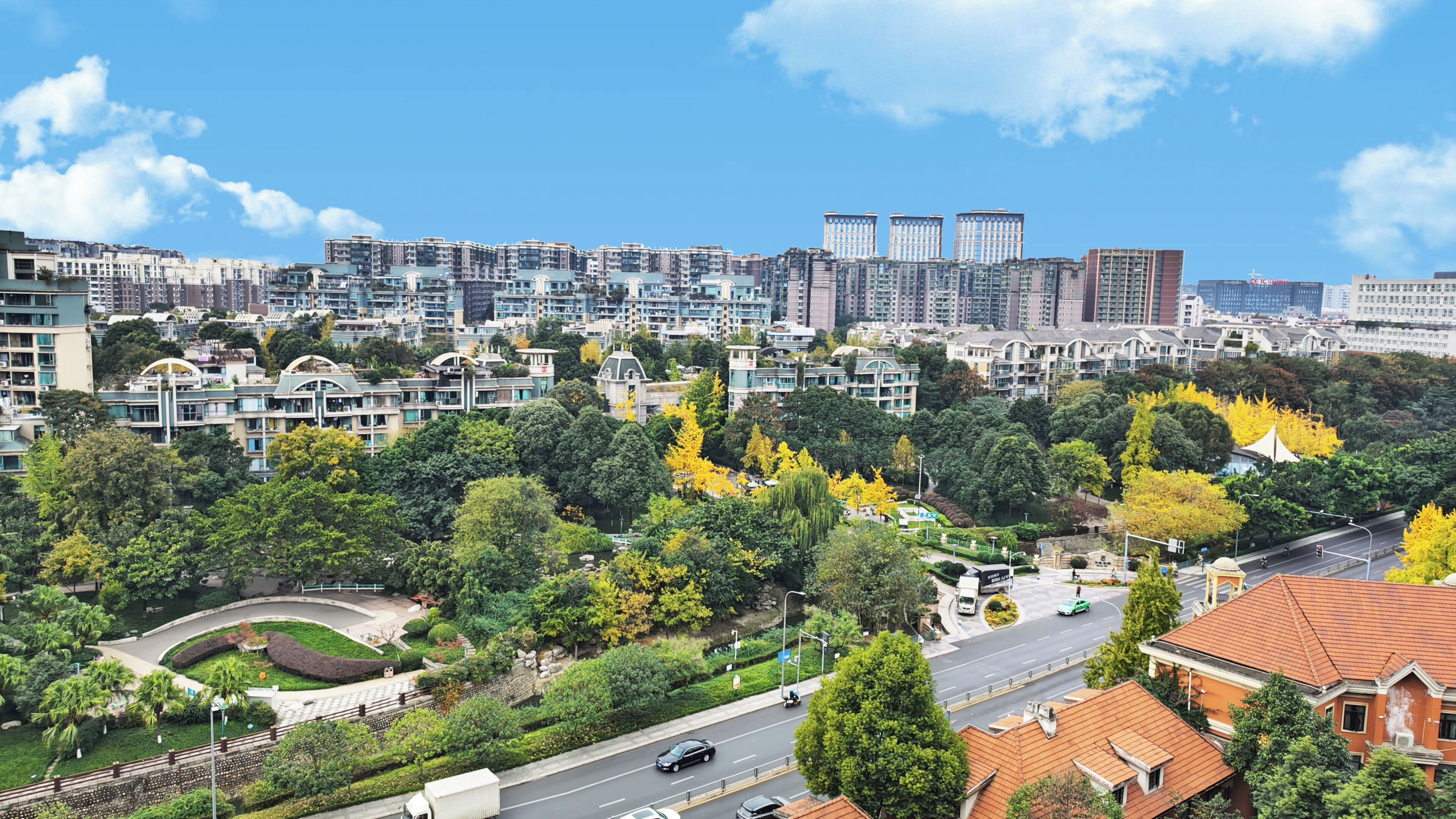 A cityscape with modern high-rise buildings, a well-maintained park with trees, and a road with vehicles.