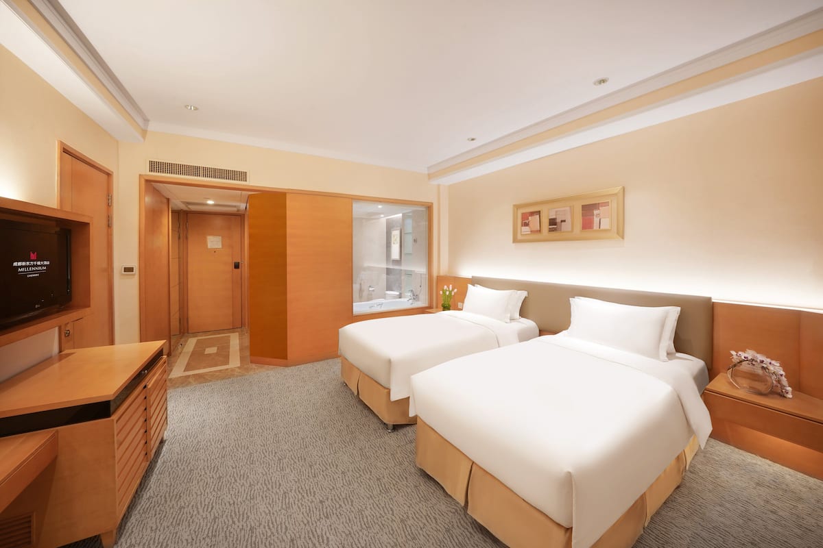 Habitación de hotel con dos camas, televisión, escritorio y baño con puerta de vidrio.