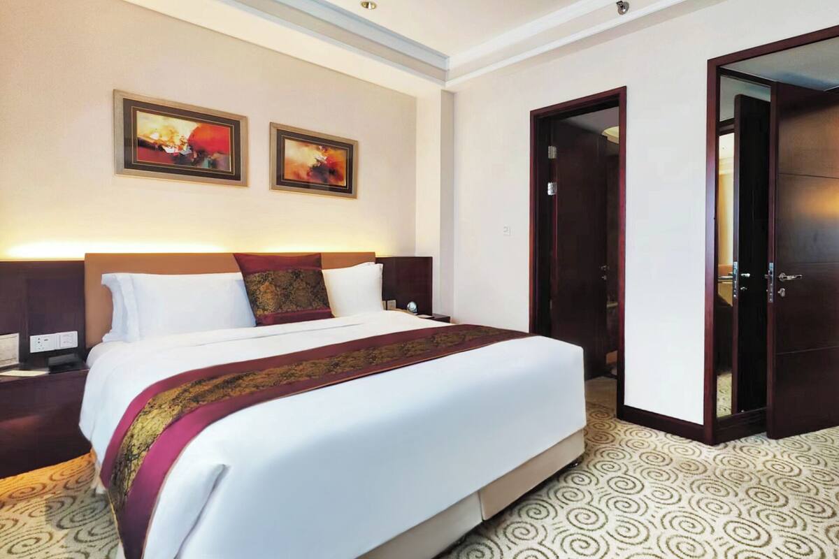 Habitación de hotel con una cama grande, dos cuadros en la pared, una mesita de noche y una puerta que da a otra habitación.