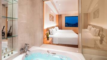 A modern hotel room with a large bed, a bathtub with rose petals, a city view, and a glass shower enclosure.