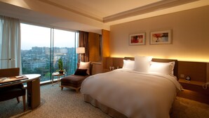 Habitación de hotel con una cama grande, un escritorio, una silla, una mesita con una planta y vistas a la ciudad a través de amplias ventanas.