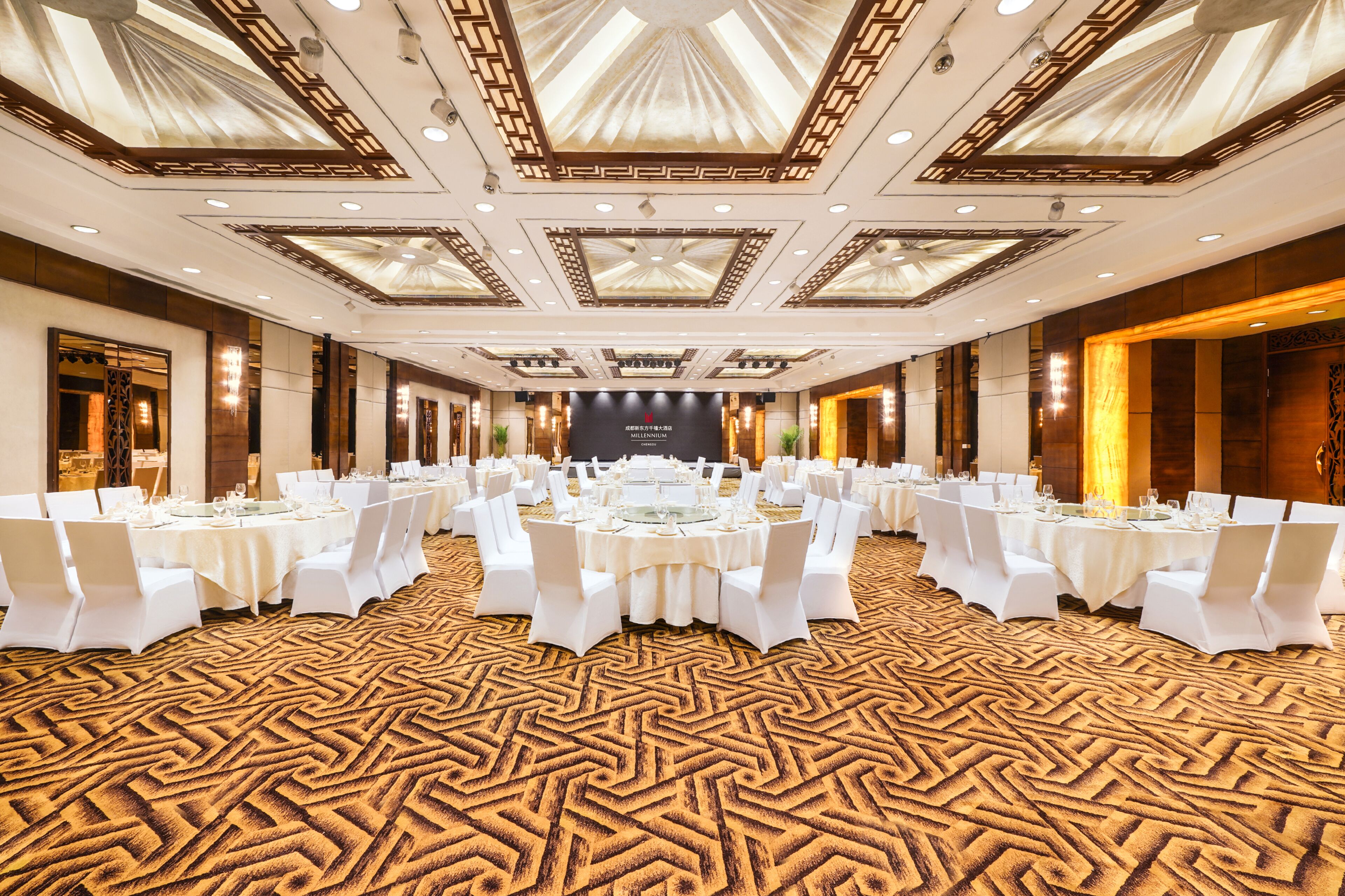 Une salle de banquet avec des tables rondes, des chaises blanches et un tapis à motifs.