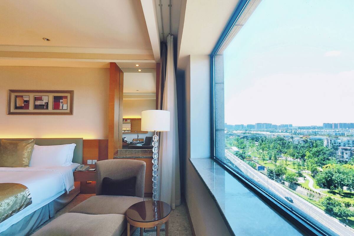 Habitación de hotel con cama, una silla, una mesita y una lámpara. Se aprecia una ventana grande con vistas a la ciudad.