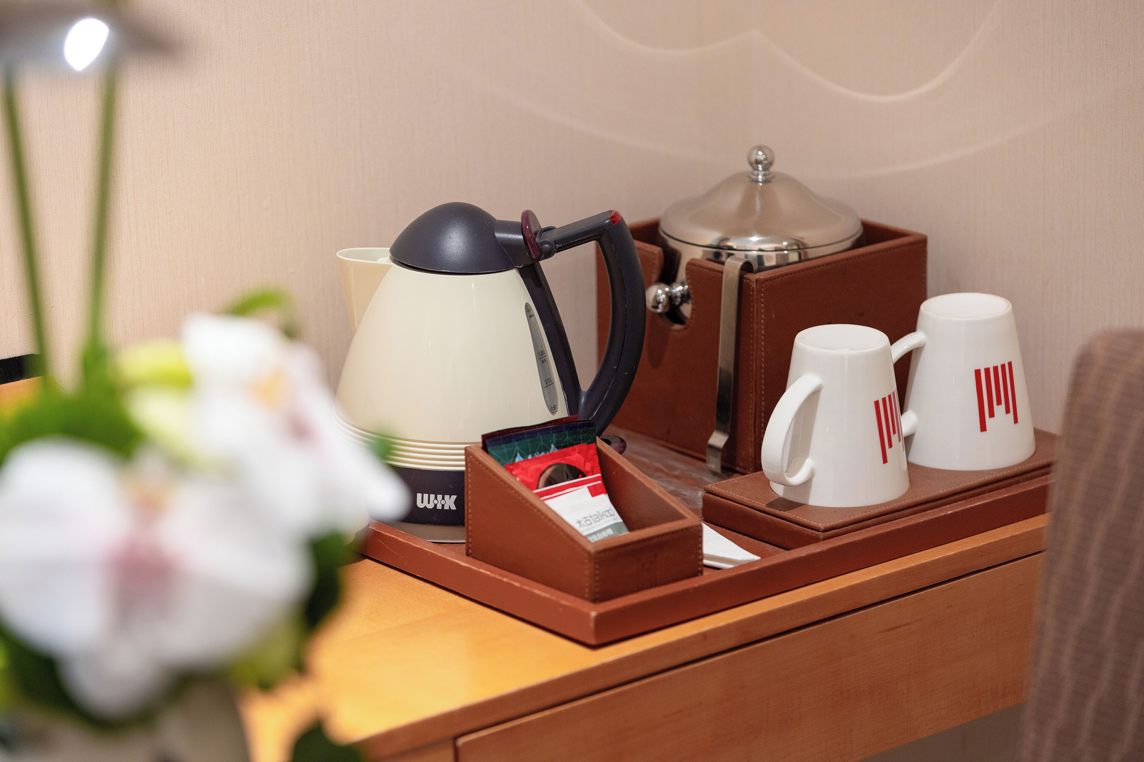 Habitación de hotel impecable, con una mesita de noche de madera, una lámpara blanca, una cafetera, dos tazas blancas y una pequeña caja de pañuelos.