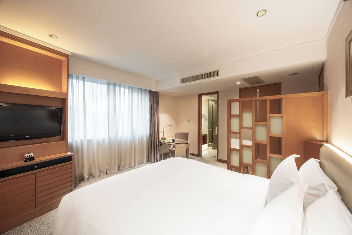 Habitación de hotel con una cama grande, un televisor sobre un mueble de madera, una mesita con una silla, y un baño con espejo y mampara de cristal.