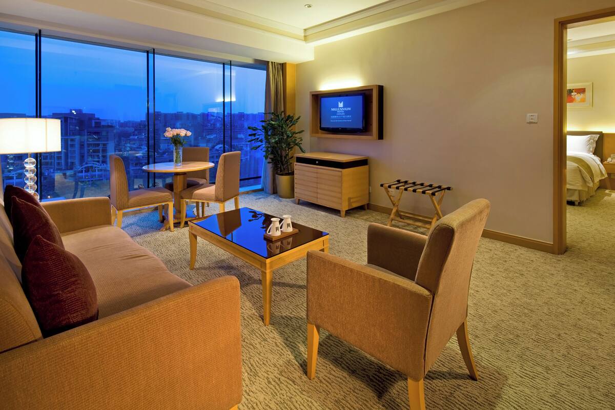 Habitación de hotel moderna con un ventanal grande que ofrece vistas a la ciudad, un televisor de pantalla plana montado en la pared, un rincón de descanso acogedor con sofá y sillones, y una pequeña mesa de comedor.