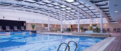 An indoor swimming pool with a glass ceiling, lounge chairs, and a wooden deck area.
