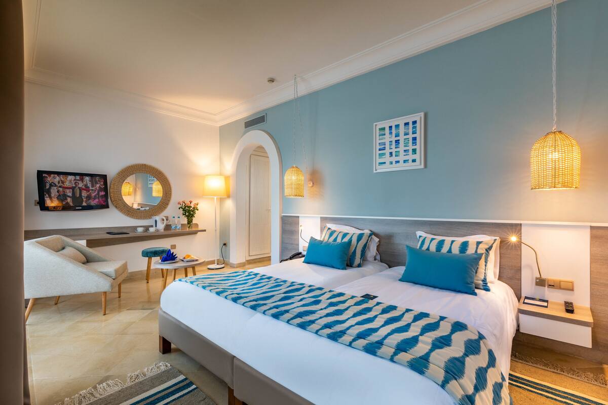 A modern hotel room with a large bed, a sitting area with a chair, a desk, and a television.