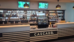 A bar area with a counter labeled 'CASCADE,' shelves with bottles and glasses, and two screens displaying content. - Hotel Cascade (Most)