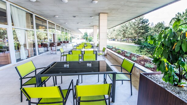 An outdoor seating area with tables and chairs, large windows, and a view of the surrounding greenery.