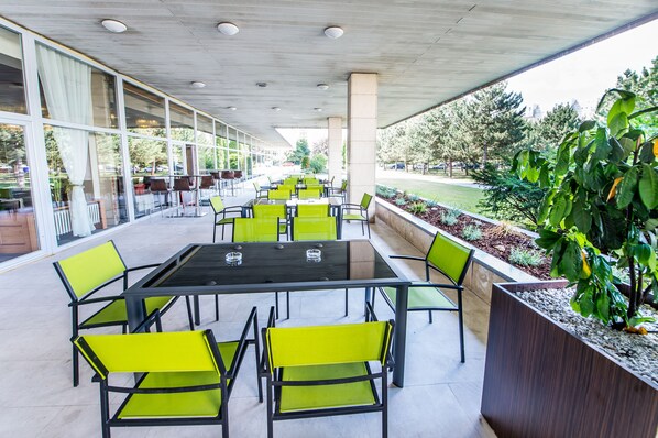 An outdoor seating area with tables and chairs, large windows, and a view of the surrounding greenery.