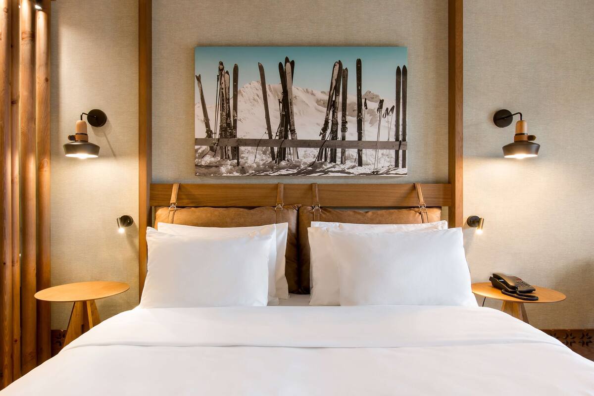 Camera d'albergo con un letto, due comodini, un quadro raffigurante sci su una montagna e lampade a parete.