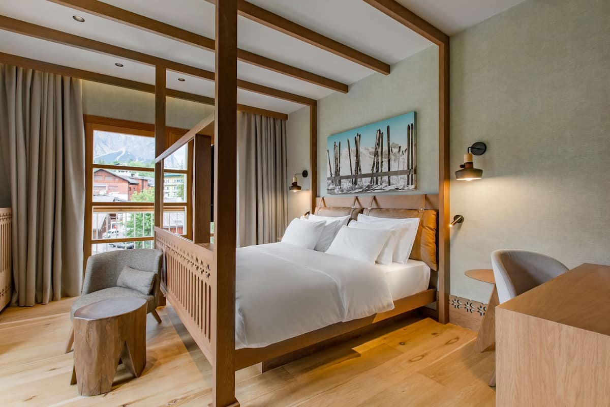 Un letto con struttura in legno e biancheria bianca, un piccolo tavolino e una sedia in una stanza con pavimento in legno e una grande finestra.