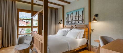 A wooden-framed bed with white bedding, a small wooden table, and a chair in a room with wooden floors and a large window.