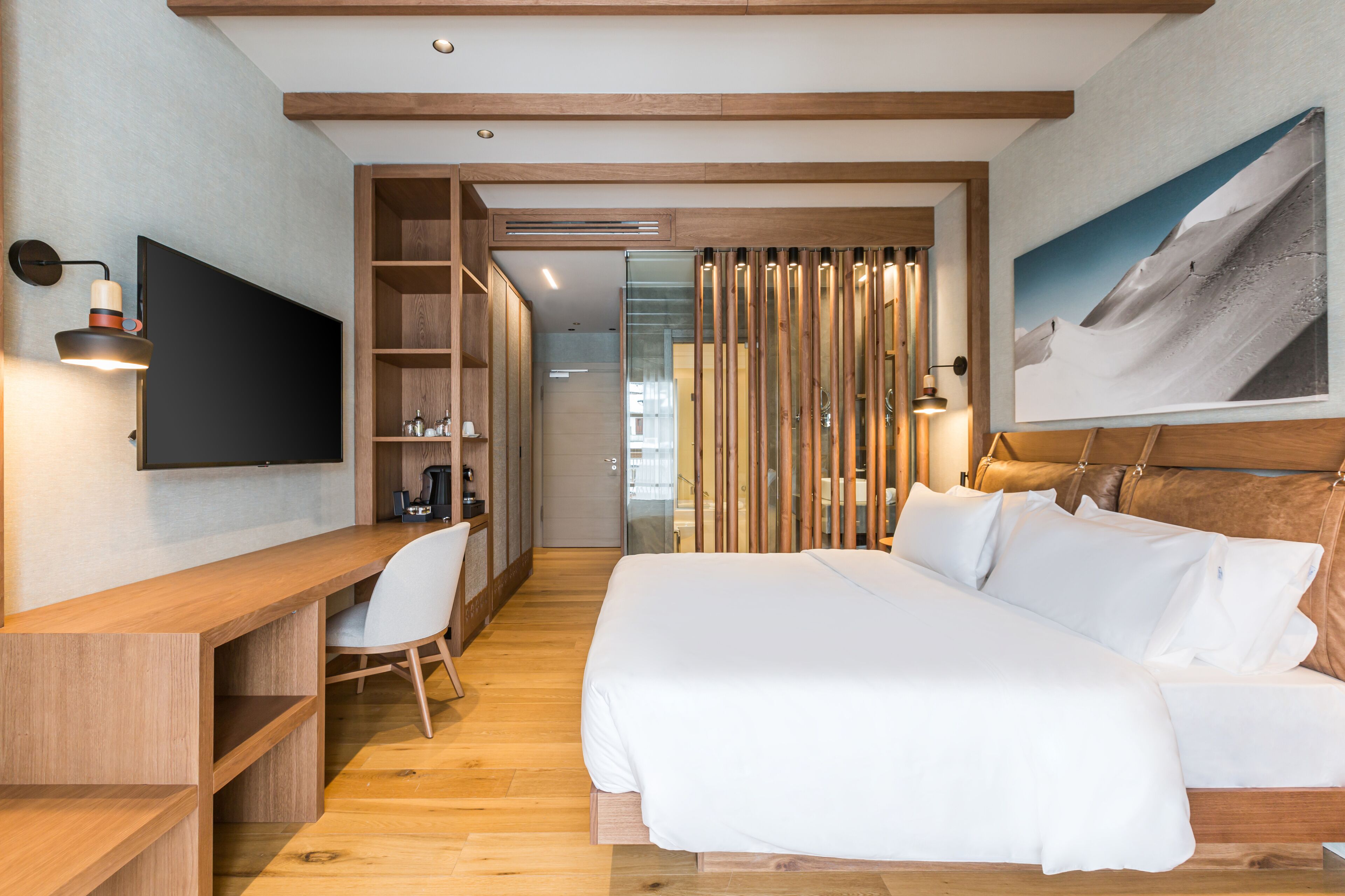 Una habitación de hotel moderna con un escritorio de madera, un televisor de pantalla plana, una cama con sábanas blancas y un ventanal con vistas a la montaña.