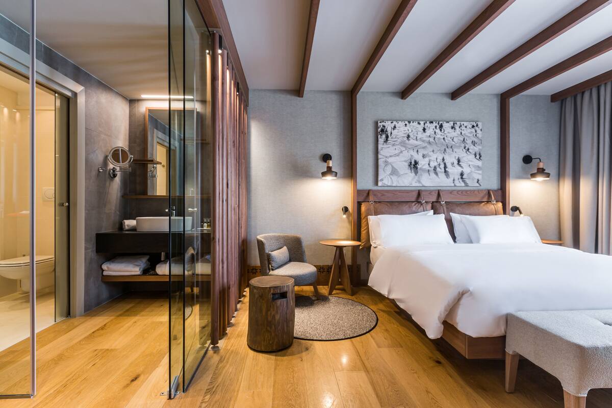 Una moderna camera d'albergo con pavimento in legno, bagno con pareti in vetro, un letto con lenzuola bianche, un tavolino rotondo in legno e un quadro appeso al muro.